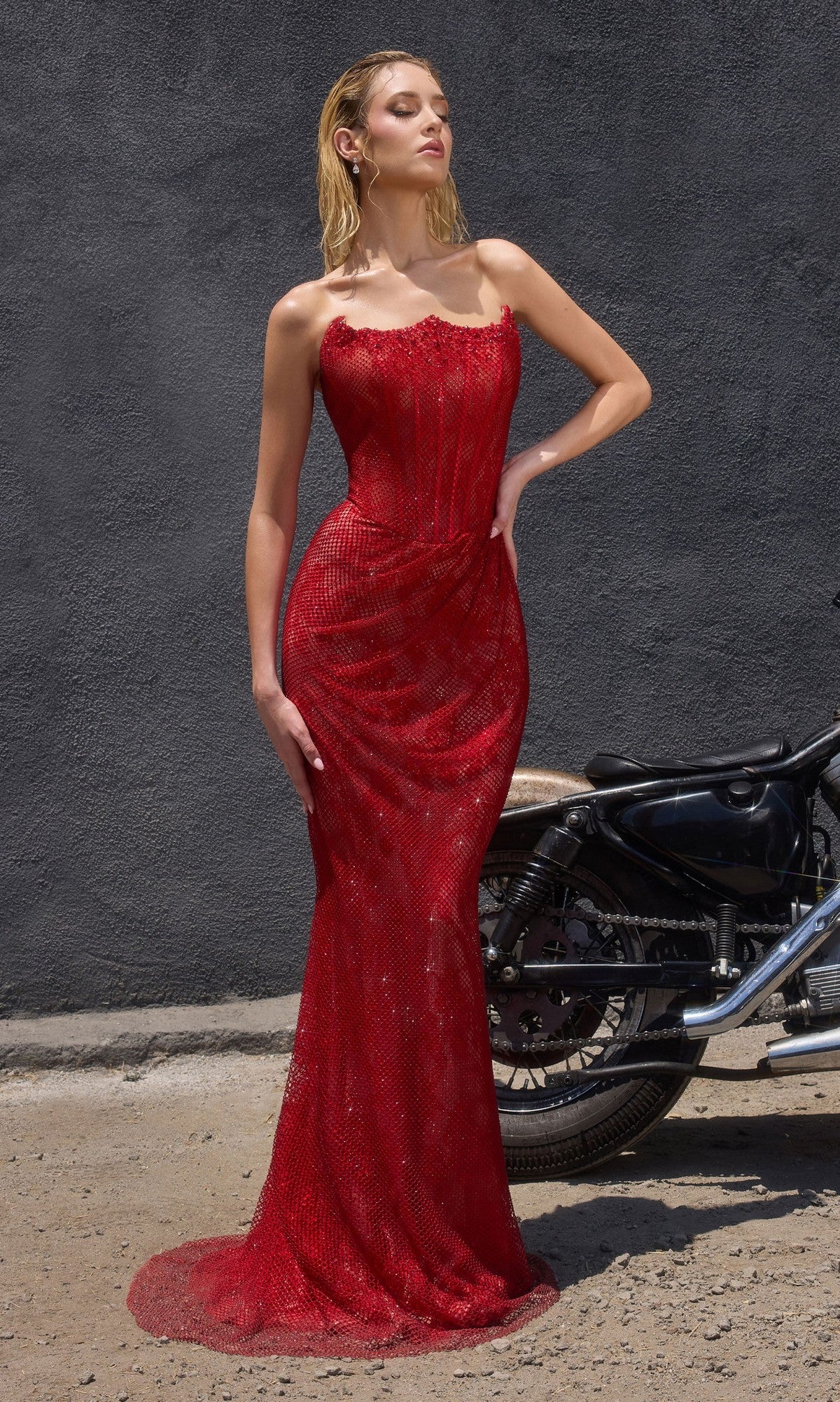 A woman in the Ladivine CB162C long plus-size prom dress, featuring a fitted corset bodice and strapless design, stands beside a black motorcycle against a dark gray wall on a sunny day.